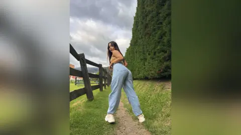 Young woman in light blue jeans and a tan top posing by a wooden fence in a grassy field under a cloudy sky.