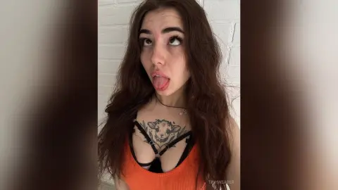 Playful young woman with long wavy hair wearing an orange tank top against a white brick wall.