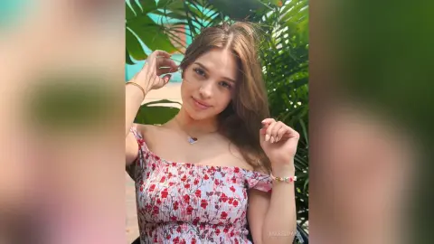 Smiling young woman in a floral off-the-shoulder top posing in a lush tropical garden setting.