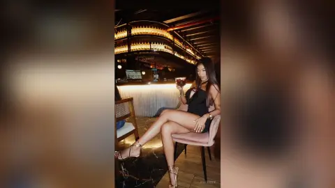 Elegant woman in a black cocktail dress sitting at a dimly lit luxury bar holding a drink.