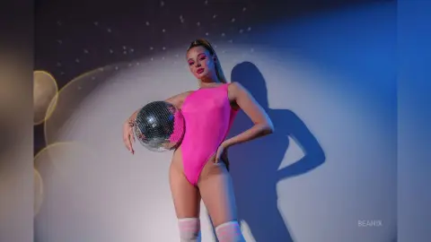 BeaNix poses in a bright pink bodysuit while holding a large disco ball against a blue-lit wall.