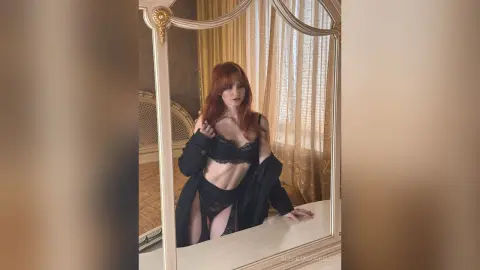 AlbertaCorrell poses in black lace lingerie and an open black coat while looking into a mirror.