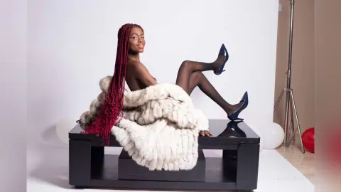 Smiling Black woman with long red braids wearing a luxury white faux fur coat sitting on a black bench.