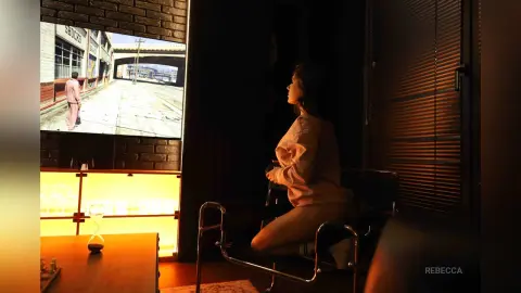 Rebecca sits in a chair in a dark room, looking toward a large screen displaying a street scene.