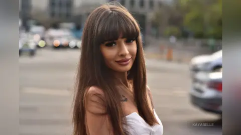 Smiling young woman with long brown hair and bangs wearing a white top on a sunny city street.