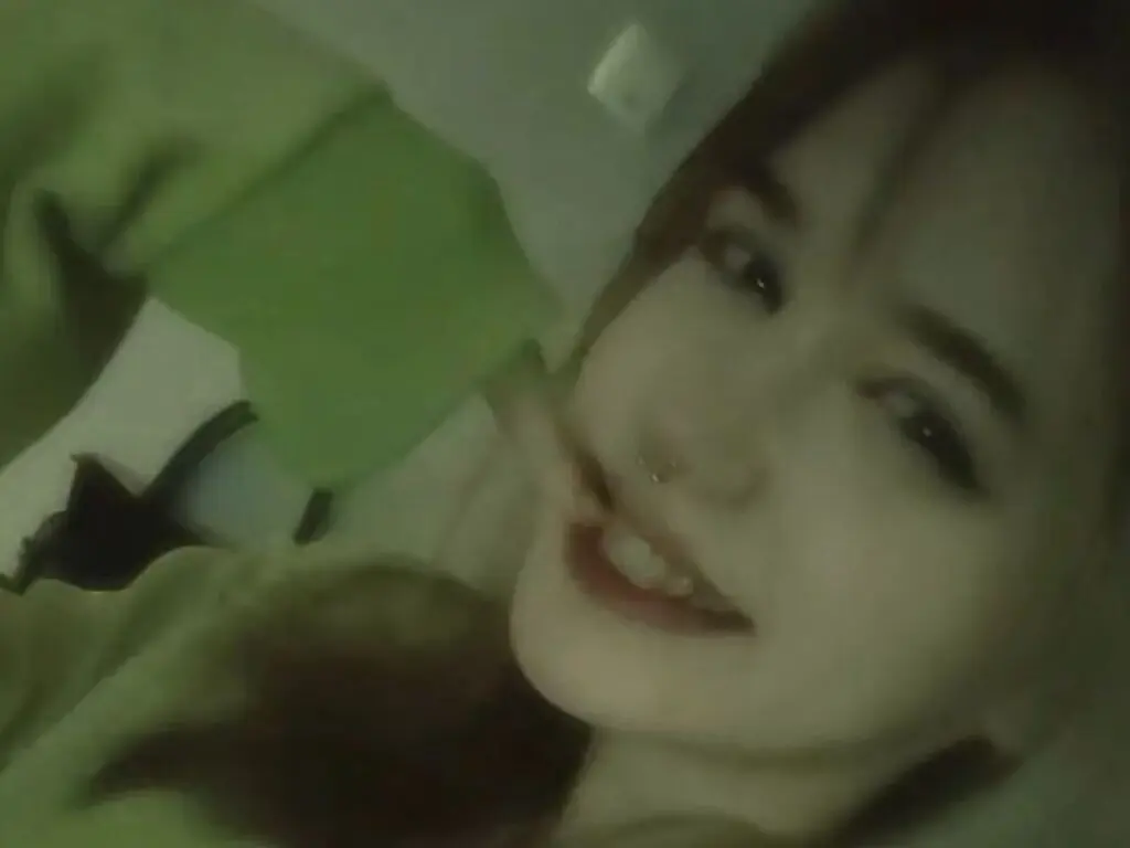 Smiling young woman in a green sweater relaxing in a cozy, dimly lit indoor setting.