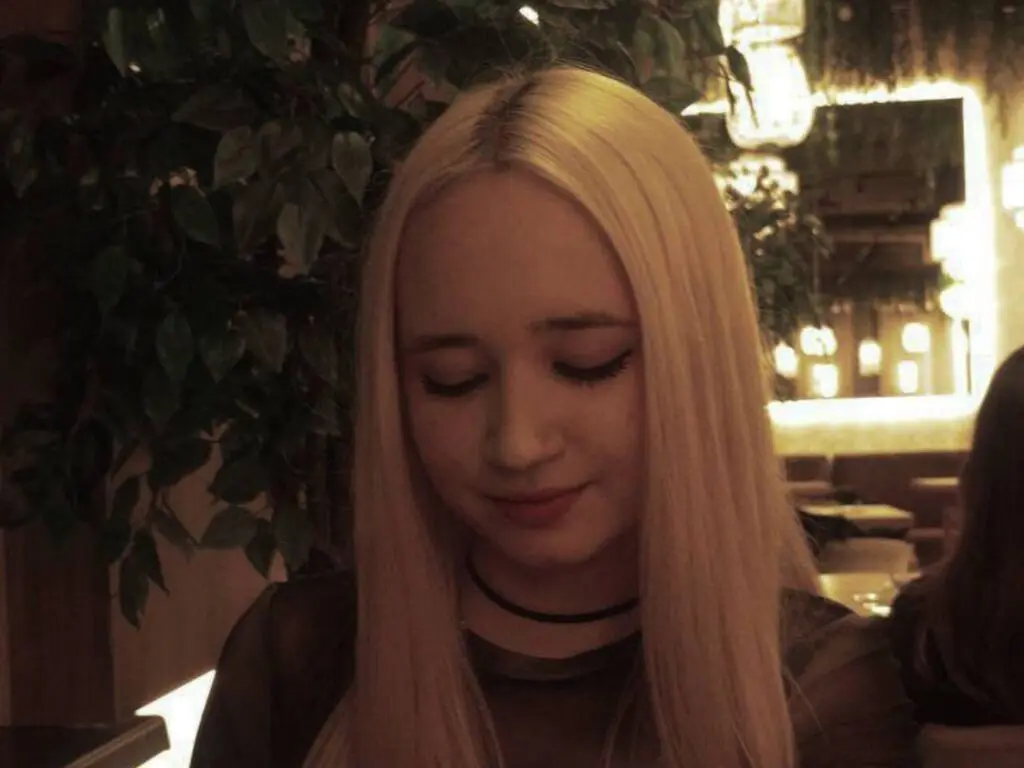 Blonde woman in a black mesh top sitting in a dimly lit restaurant with lush green plants.