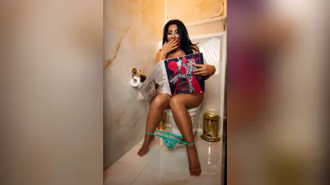 MissyJolie sits on a white toilet wearing light blue lace lingerie while holding an open magazine in front of her chest.