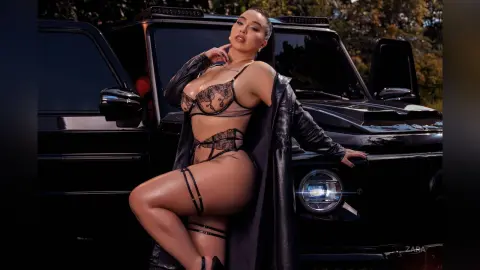 Curvy woman in black lace lingerie and leather jacket posing confidently in front of a black SUV outdoors.