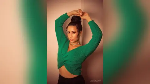 Young woman in a green knit crop top posing stylishly against a neutral wall in soft lighting.