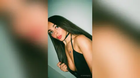 Young woman with long dark hair wearing a black halter top in a minimalist indoor setting with soft lighting.