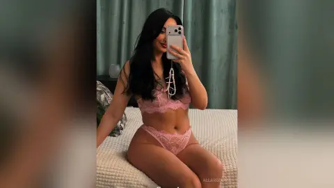 Young woman in pink lace lingerie taking a mirror selfie in a bright bedroom setting.