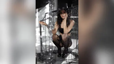 Elegant woman in black lingerie posing behind shimmering silver beaded curtains in a moody, cinematic setting.