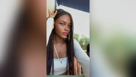 Young woman with long dark braids wearing a white long-sleeve top in a bright outdoor setting.