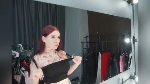 Red-haired woman in a black crop top styling outfits in a modern walk-in closet with clothing racks.