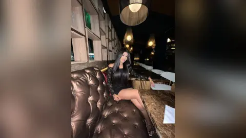 Elegant woman in a black long-sleeve dress sitting on a luxury leather booth in a moody, dimly lit restaurant.