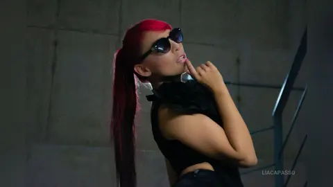 Stylish woman with pink hair and sunglasses posing in a black sleeveless top against a moody urban background.
