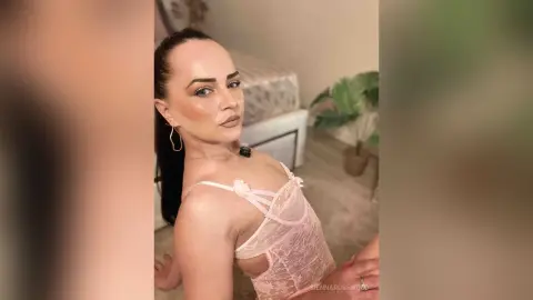 Glamorous woman in a pink lace bodysuit posing in a bright, modern bedroom setting.