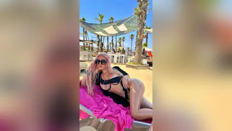 Stylish woman in a black bikini and sunglasses relaxing on a pink towel at a sunny tropical beach resort.