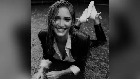 TiffanyAddams smiles while lying in shallow water, wearing a white collared shirt and dark pants.