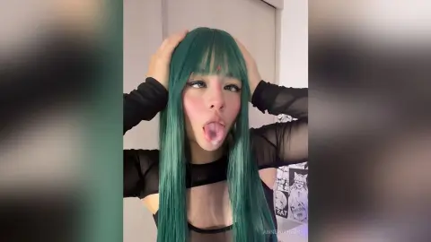 Young woman with long green hair and black mesh top posing playfully in a bright indoor setting.