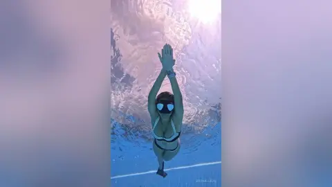 Young woman in a white bikini swimming underwater in a bright blue pool with sunlight reflecting through the water.