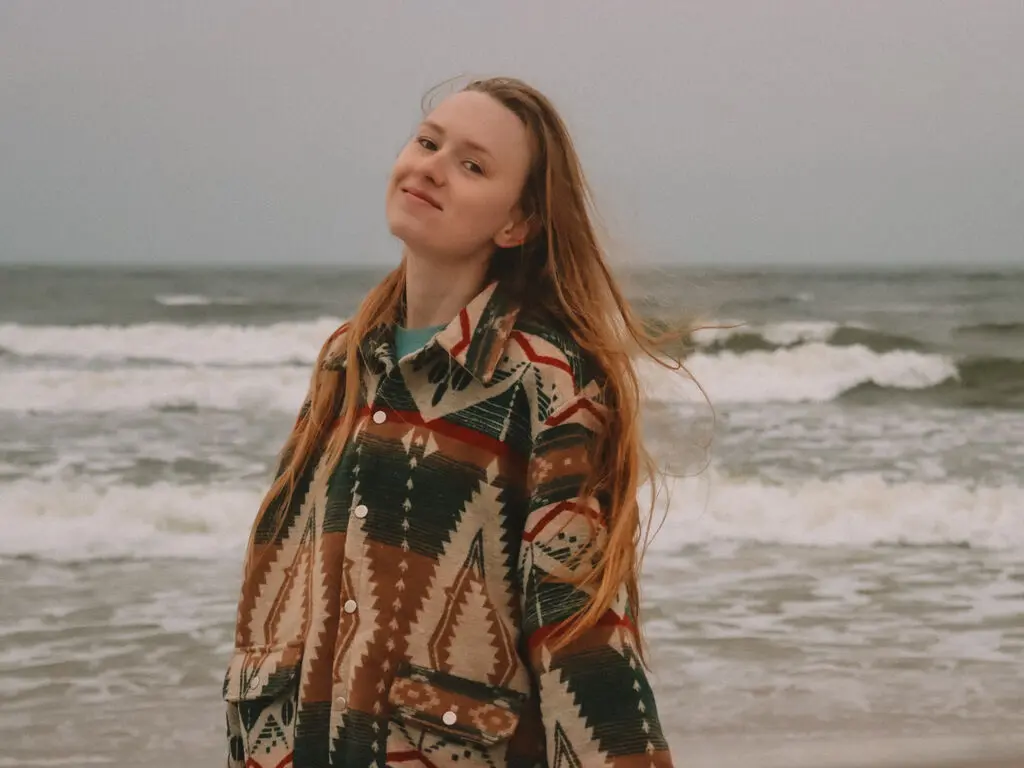 Smiling woman in a patterned bohemian jacket standing on a windy beach under a cloudy sky.