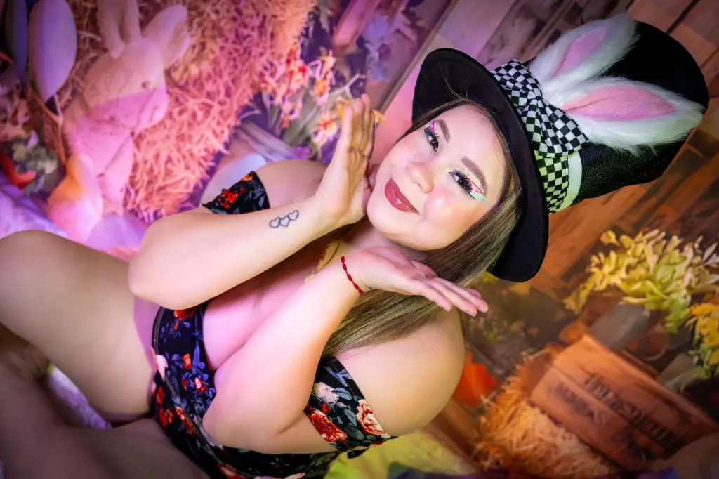 katheringoezz69 poses with her blonde hair styled up, wearing a black sleeveless top against a decorative floral and patterned background.