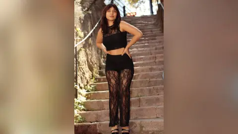 Stylish woman in a black lace crop top and sheer lace trousers posing on stone stairs in soft sunlight.
