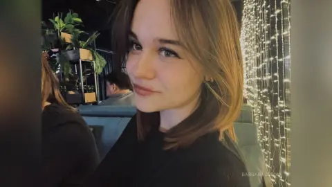 Smiling young woman in a black top posing in a cozy cafe with warm fairy lights in the background.