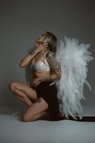 Tattooed woman in white angel wings and sparkly bikini top posing gracefully against a grey studio background.