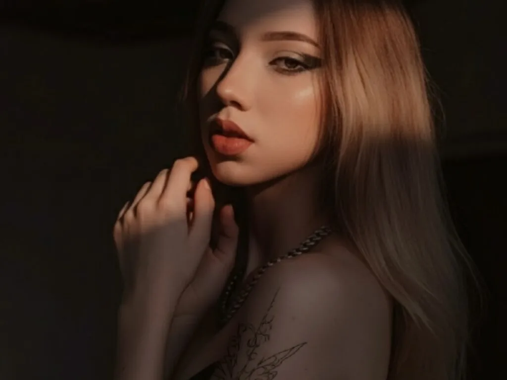 ValkaRosselli — ValkaRosselli poses in low lighting with long blonde hair and a silver chain necklace.