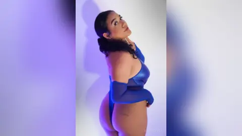 Confident woman in a blue bodysuit posing against a soft purple studio background.