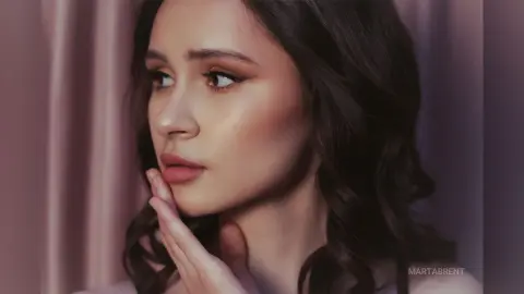 Young woman with glowing makeup and wavy hair looking thoughtfully in a soft, moody indoor setting.