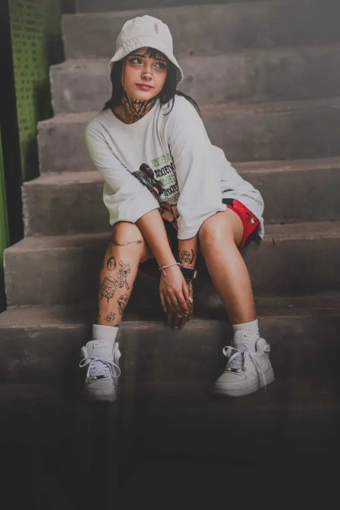 Tattooed woman in a white crop top and blue skirt sitting on a newspaper background with blue lighting.