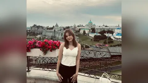 Young woman in a white tank top standing on a balcony overlooking a historic European city skyline at dusk.