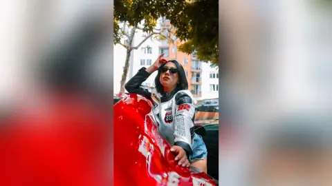 KimRoberth poses in a black and white leather jacket and denim shorts while leaning against a bright red car outdoors.