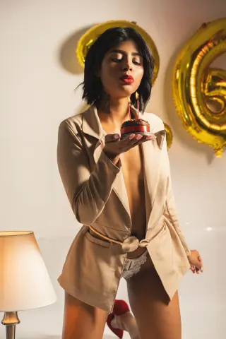 Elegant woman in a beige blazer and lace lingerie holding a chocolate cake near gold birthday balloons.