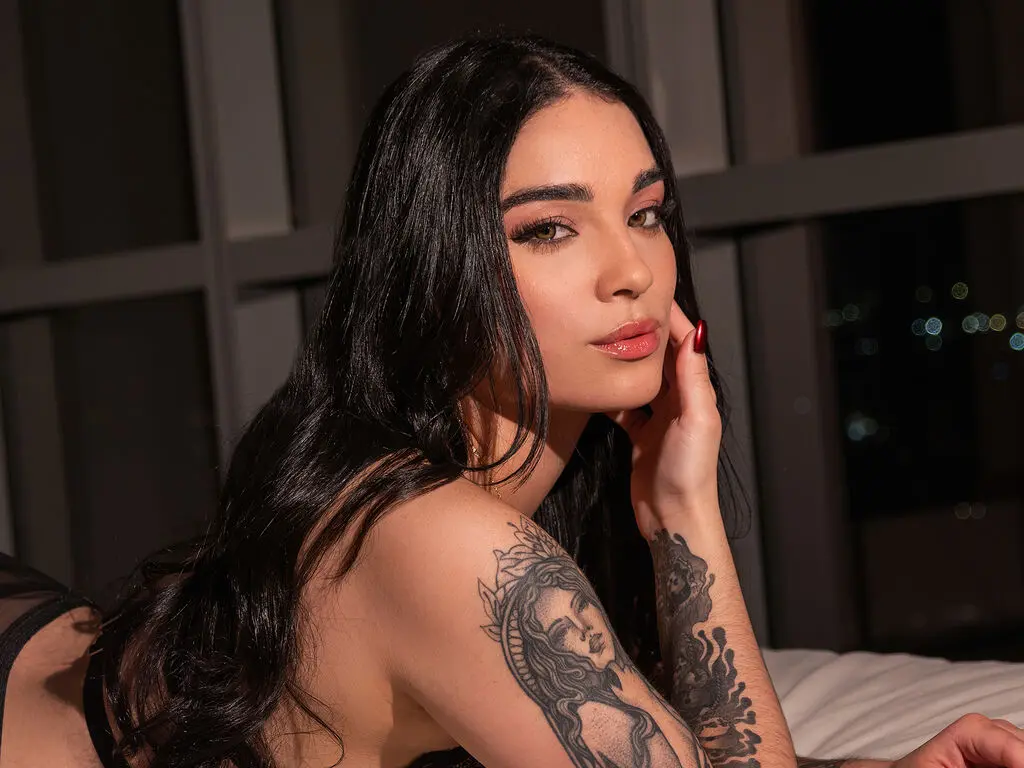 AdalineCruz poses with long black hair and visible tattoos on her shoulder and hand, looking over her shoulder while resting on a bed.