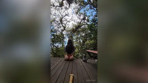 RossiNoleen sits on a wooden deck outdoors, showing her bare buttocks and back while facing away from the camera toward the trees.
