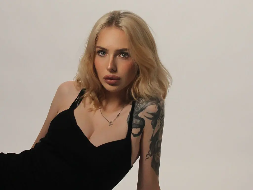 Blonde woman in a black tank top with arm tattoos posing against a neutral studio background.