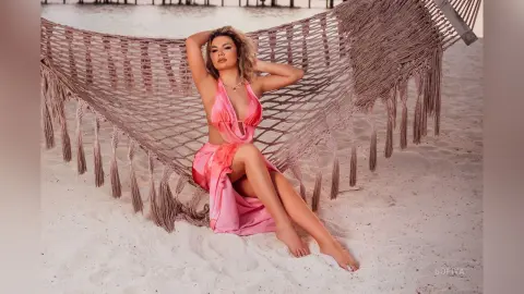 Relaxed woman in a pink summer dress lounging in a large macrame hammock on a sunny beach.