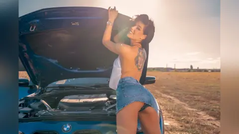 Smiling woman in a white tank top and denim mini skirt posing by a blue car in a sunny field.