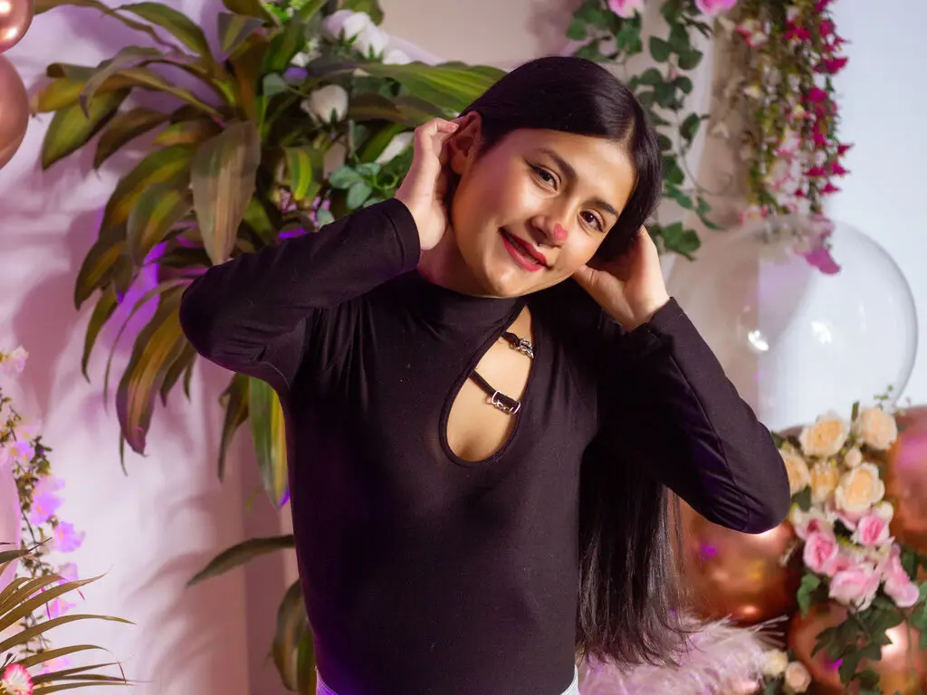 Smiling young woman in a black long-sleeve top posing amidst pink flowers and tropical greenery.