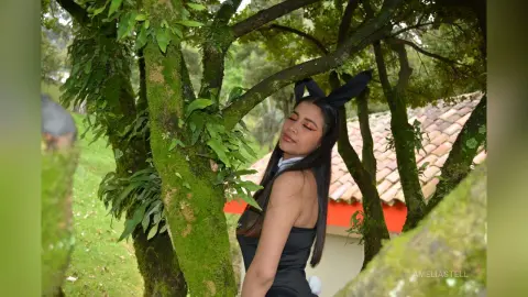AmeliaStell poses outdoors near a mossy tree trunk wearing a black bunny ear headband and a black top.