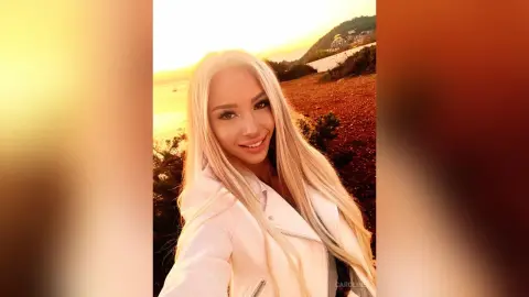 Smiling blonde woman in a white jacket enjoying a golden sunset on a coastal hillside.