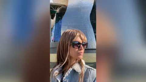 Stylish woman with long blonde hair wearing sunglasses and a denim jacket in front of modern architecture.