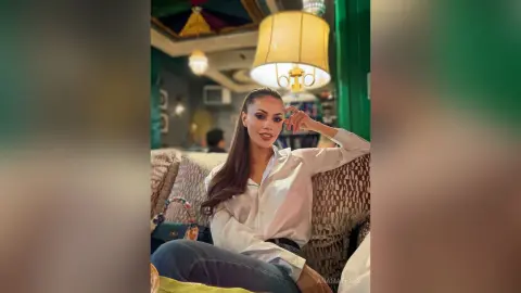AnaMathers sits on a patterned sofa wearing a white button-down shirt and blue jeans, resting her hand near her face.