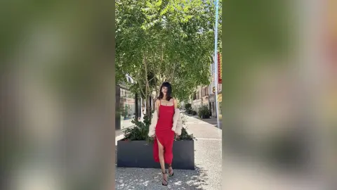 Stylish woman in a red midi dress and white faux fur coat walking on a sunny city street lined with green trees.