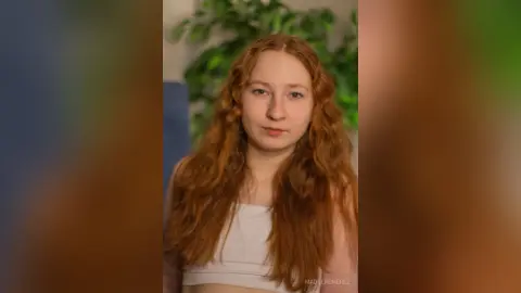 Redheaded woman in a white crop top posing in a bright indoor setting with green plants.